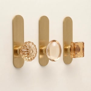 May include: Three wall-mounted hooks featuring vertical brushed brass rectangular backplates. Each hook is topped with a distinct glass knob: one round and fluted, one smooth and oval, and one square with a clear, faceted amber-toned finish.