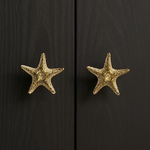 K&ouml;nnte beinhalten: Zwei goldfarbene Seestern-Schrankkn&ouml;pfe vor einem dunklen Holzgrund. Die Kn&ouml;pfe haben strukturierte Oberfl&auml;chen und verleihen einen Hauch von K&uuml;sten-Dekor. Die Kn&ouml;pfe sind an einem dunklen Holzschrank befestigt.