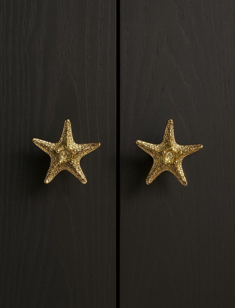 K&ouml;nnte beinhalten: Zwei goldfarbene Seestern-Schrankkn&ouml;pfe vor einem dunklen Holzgrund. Die Kn&ouml;pfe haben strukturierte Oberfl&auml;chen und verleihen einen Hauch von K&uuml;sten-Dekor. Die Kn&ouml;pfe sind an einem dunklen Holzschrank befestigt.