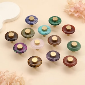May include: A collection of decorative cabinet knobs in various colors and patterns. Each knob features a round, polished stone top with a brass-colored center and stem. The knobs are in shades of blue, green, red, purple, and brown.