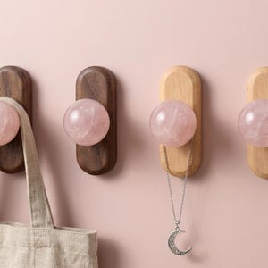 Può includere: Quattro ganci da parete con pietre di quarzo rosa sferiche e levigate montate su basi rettangolari in legno. Due ganci sono in legno scuro e due in legno chiaro. Una borsa in tessuto beige e una collana d'argento sono appese ai ganci.