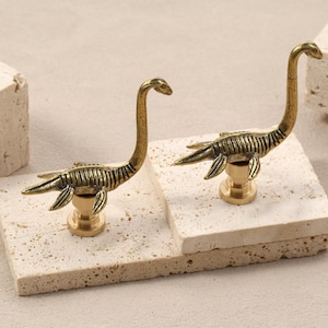 Peut inclure: Deux figurines de dinosaures en laiton avec de longs cous et des appendices en forme de nageoires. Les figurines sont montées sur des bases dorées et reposent sur des dalles de pierre de couleur claire. Les dinosaures sont détaillés avec des corps nervurés.
