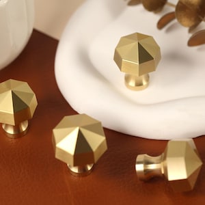 May include: Four gold-colored metal cabinet knobs with a faceted, octagonal top design. Each knob features a smooth, brushed finish and a central point that slopes down to eight distinct edges, creating a geometric shape for furniture hardware.