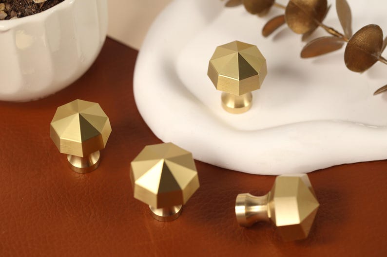 May include: Four gold-colored metal cabinet knobs with a faceted, octagonal top design. Each knob features a smooth, brushed finish and a central point that slopes down to eight distinct edges, creating a geometric shape for furniture hardware.