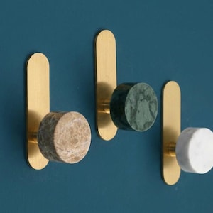 Puede incluir: Tres ganchos de pared con placas verticales de latón cepillado y pomos cilíndricos. Los pomos están hechos de piedra natural en tres colores distintos: una piedra de mármol beige, una piedra de mármol verde oscuro y una piedra de mármol blanca.