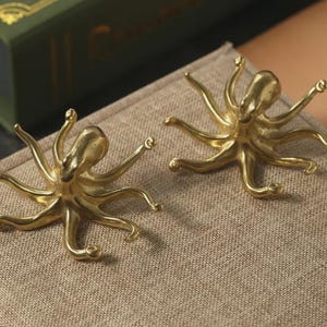 Könnte beinhalten: Zwei goldfarbene, tintenfischförmige Dekorationsgegenstände. Jeder Tintenfisch hat acht Tentakel und einen abgerundeten Kopf. Sie sind auf einer strukturierten, neutral getönten Oberfläche platziert. Die Objekte scheinen aus Metall zu sein und sind sehr detailliert.