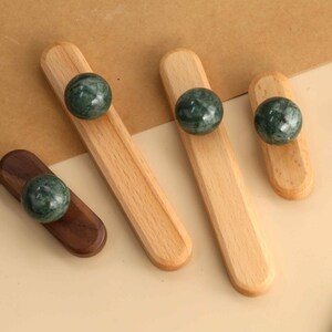 Puede incluir: Cuatro tiradores de armario, cada uno con un pomo de mármol verde oscuro, liso y redondo, unido a una base de madera rectangular y alargada. Tres bases son de madera clara, mientras que una es de madera marrón oscura con extremos redondeados.