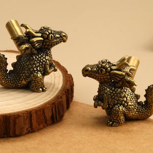 May include: Two antique brass-toned metal drawer knobs shaped like small dragons. Each dragon figure features detailed scales, horns, and a curved tail, with a cylindrical mounting post extending from the back of the creature's body.