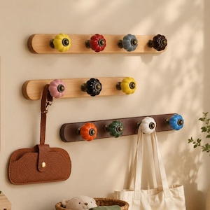 Op de afbeelding: Houten kapstokken met kleurrijke keramische knoppen. De rekken zijn gemonteerd op een beige muur, met een bruin leren etui en een canvas draagtas die aan de knoppen hangen. De knoppen zijn geel, rood, blauw en bruin.