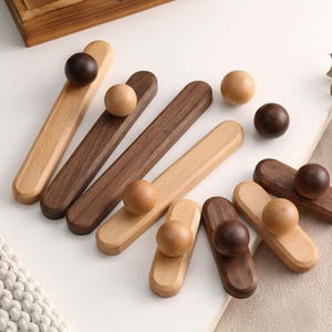 Op de afbeelding: Een verzameling houten kastgrepen in verschillende tinten bruin en beige. Elke greep heeft een afgeronde knop die aan een rechthoekige basis is bevestigd. De grepen zijn gerangschikt op een wit oppervlak, waarbij de verschillende houtnerven te zien zijn.