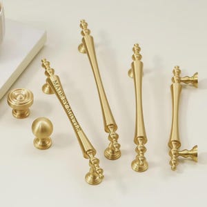 Può includere: Una collezione di ferramenta dorata per armadi, tra cui pomelli e maniglie. Le maniglie variano in lunghezza e design, con dettagli ornati. Una maniglia ha il testo "MADE BY MRVILLE". La ferramenta è disposta su una superficie bianca.