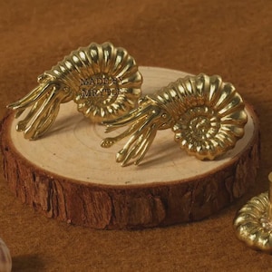 May include: Two brass cabinet knobs in the shape of ammonite fossils, with a spiral design and tentacle-like protrusions. The knobs are displayed on a wooden slice, with seashells nearby. The text "MADE BY MRYTIE" is visible.