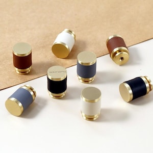May include: A collection of cylindrical cabinet knobs with brass-colored tops and bottoms. The knobs have leather or painted bands in various colors, including brown, white, black, and blue. The knobs are arranged on a neutral background.