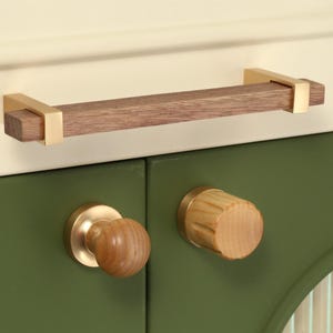 Könnte beinhalten: Schrankbeschlagsatz mit einem Holzgriff, ca. 15 cm lang, und goldfarbenen Endkappen sowie zwei Holzknöpfen mit goldfarbenen Basen. Montiert an einer grünen Schranktür.