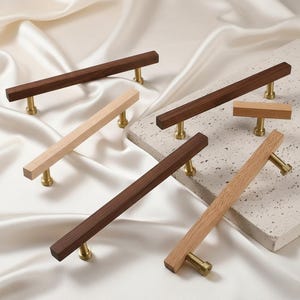 May include: A collection of wooden cabinet pulls in various shades of brown and natural wood. Each pull features a rectangular wooden handle and brass-colored hardware. The pulls are arranged on a white surface and a textured, light gray surface.