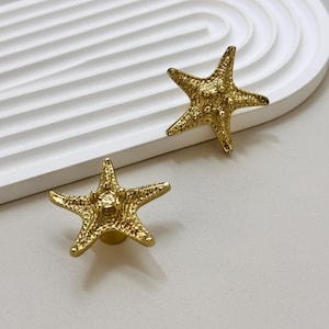 K&ouml;nnte beinhalten: Zwei goldfarbene, sternf&ouml;rmige Schrankkn&ouml;pfe. Die Kn&ouml;pfe haben eine strukturierte Oberfl&auml;che und sind auf einer kleinen, runden Basis montiert. Die Kn&ouml;pfe werden auf einer wei&szlig;en Oberfl&auml;che pr&auml;sentiert.