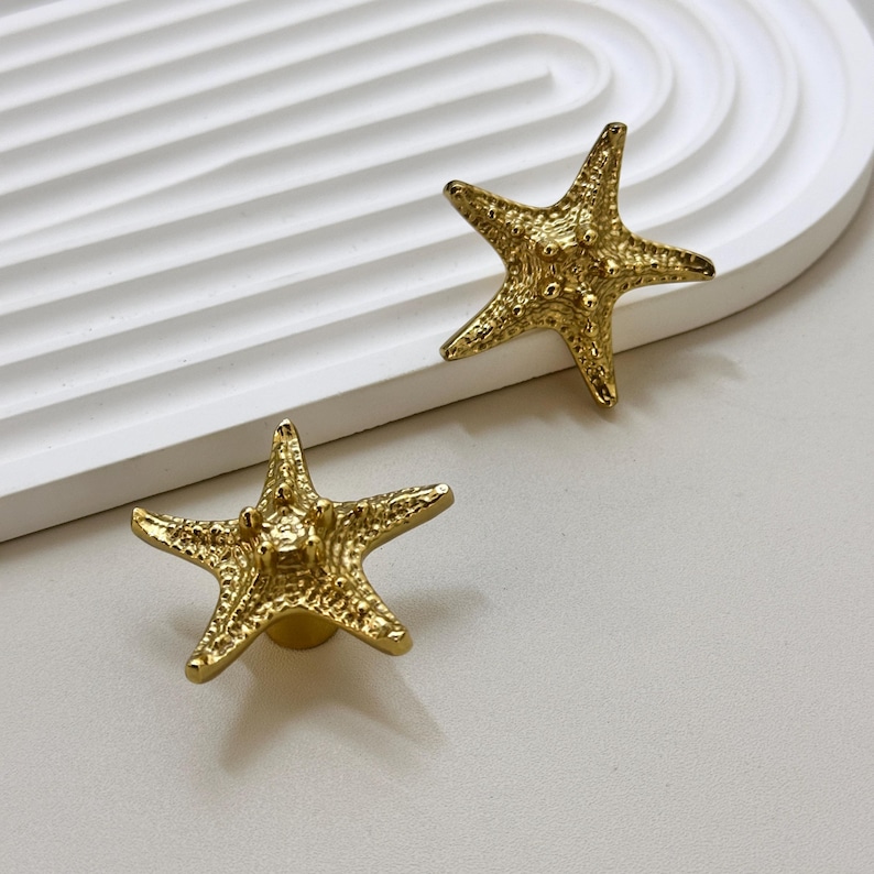 K&ouml;nnte beinhalten: Zwei goldfarbene, sternf&ouml;rmige Schrankkn&ouml;pfe. Die Kn&ouml;pfe haben eine strukturierte Oberfl&auml;che und sind auf einer kleinen, runden Basis montiert. Die Kn&ouml;pfe werden auf einer wei&szlig;en Oberfl&auml;che pr&auml;sentiert.