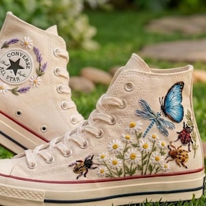 Könnte beinhalten: Cremefarbene High-Top-Sneaker mit gestickten Blumen- und Insektenmotiven. Die Sneaker haben einen Converse All Star Logo-Patch und einen roten Streifen auf der Sohle. Die Stickereien zeigen Gänseblümchen, einen blauen Schmetterling und Käfer.