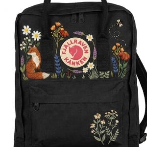 May include: A black backpack featuring a circular Fjällräven Kånken logo patch on the front. The bag is decorated with embroidered floral patterns, a small fox on the left side, and a tiny bee on the upper right. It includes a front zippered pocket.