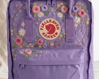 Può includere: Uno zaino lavanda con ricami floreali, tra cui rose rosa, margherite bianche e dettagli gialli. Lo zaino presenta una tasca frontale, una maniglia superiore e il logo Fjallraven Kanken.