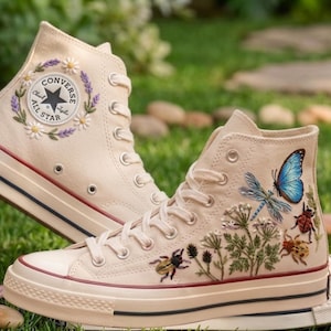 Könnte beinhalten: Ein Paar cremefarbene High-Top-Sneaker aus Segeltuch mit detaillierten Stickereien. Ein Schuh zeigt einen Blumenkranz um das Converse All Star-Logo, während der andere einen blauen Schmetterling, eine Libelle und verschiedene Käfer darstellt.