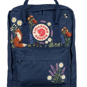 May include: A navy blue backpack featuring an embroidered design of a fox, various wildflowers, and a small bee. The circular Fjällräven Kånken logo is centred on the front. The bag includes a top handle, two shoulder straps, and a front zipper pocket.