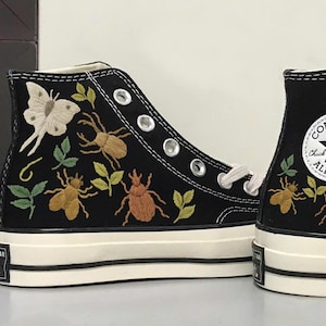 Könnte beinhalten: Schwarze High-Top-Sneaker mit gestickten Insekten- und Blattmotiven. Die Sneaker haben eine weiße Sohle und Schnürsenkel, mit dem Converse All Star-Logo an der Seite. Zu den gestickten Insekten gehören Käfer und ein Schmetterling.