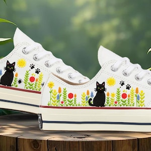 May include: White canvas trainers with a whimsical design featuring a black cat, colourful flowers, a yellow sun, and paw prints. The shoes have white laces and a cream-coloured sole with a red and navy blue stripe.
