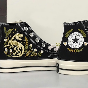 Puede incluir: Zapatillas altas negras con suelas y cordones blancos. Una zapatilla presenta un diseño de esqueleto de dinosaurio con detalles florales. La otra zapatilla tiene un logotipo de Converse All Star con detalles florales y de hojas.