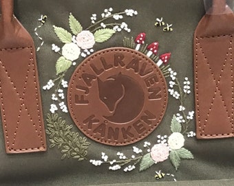 Op de afbeelding: Een groene Fjällräven Kånken rugzak met een bruine leren patch en een geborduurd bloemenontwerp. De borduurwerk bevat witte bloemen, groene bladeren en rode paddenstoelen.