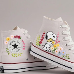 Puede incluir: Par de zapatillas altas de lona en color gris claro. Una zapatilla presenta un diseño de Snoopy con flores coloridas, mientras que la otra tiene una corona floral alrededor del logotipo de Converse All Star. Las zapatillas tienen cordones blancos y una raya roja en la suela.