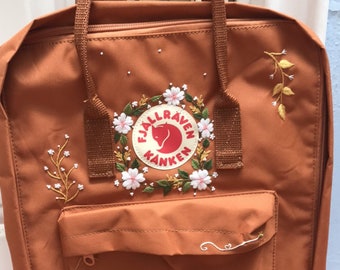Puede incluir: Una mochila Fjällräven Kånken marrón con flores y hojas bordadas alrededor del logotipo. La mochila tiene un pequeño bolsillo frontal con cremallera.