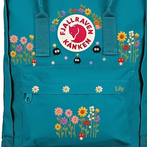 Puede incluir: Mochila turquesa con diseños bordados de flores y setas. Presenta el logotipo de la marca y el nombre "Lily" en el bolsillo delantero. Incluye asa superior y correas ajustables.