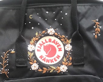 Op de afbeelding: Zwarte Fjällräven Kånken rugzak met geborduurde bloemen, bladeren en bijen. Het logo is een rode vos op een witte cirkel met de tekst "Fjällräven Kånken" in zwart.