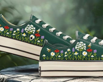 May include: A pair of dark green canvas sneakers with white laces and soles. The sneakers are decorated with embroidered flowers, bees, and other plants in various colors. The shoes are sitting on a stone surface.