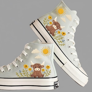 May include: Light blue high-top sneakers with embroidered designs. Each shoe features a brown Highland cow, yellow sunflowers, a yellow sun, and white clouds. The sneakers have white laces, a white sole, and a black stripe.