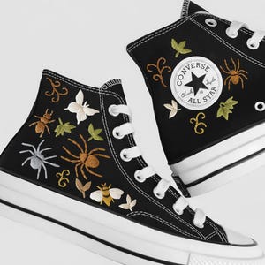 Könnte beinhalten: Schwarze High-Top-Sneaker mit weißen Schnürsenkeln und Sohlen. Die Sneaker sind mit Insekten, Blättern und Wirbeln in Braun-, Gelb- und Weißtönen bestickt. Das Converse All Star-Logo ist auf der Seite sichtbar.