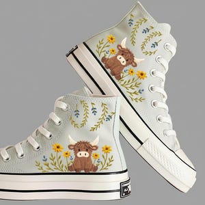 Puede incluir: Zapatillas altas de color azul claro con bordados de vaca de las Tierras Altas y diseños florales. Las zapatillas tienen una suela de goma blanca, puntera y cordones. El diseño bordado incluye una vaca marrón, flores amarillas y follaje verde y azul.