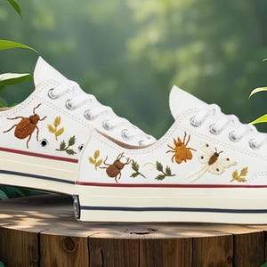 Könnte beinhalten: Ein Paar weiße Low-Top-Sneaker aus Segeltuch mit detaillierten Stickereien von braunen Käfern, grünen Blättern und einer weißen Motte. Die Schuhe haben weiße Schnürsenkel, silberne Ösen und eine dicke Gummisohle mit einem roten und blauen Streifen.