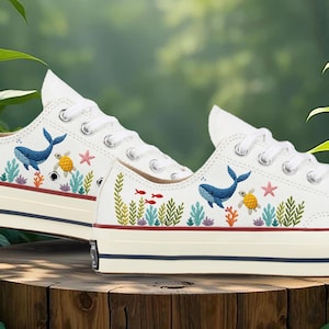 May include: White canvas trainers with embroidered aquatic designs. The trainers feature a whale, turtle, starfish, and coral reef elements. The shoes have white laces and a cream-coloured sole with a navy blue stripe.