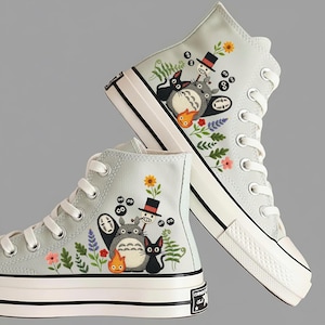 Könnte beinhalten: Hellgraue High-Top-Sneaker mit gestickten Cartoon-Charakteren und floralen Designs. Die Sneaker haben weiße Schnürsenkel, eine weiße Gummisohle und einen schwarzen Streifen. Das Design umfasst Charaktere, Blumen und Blätter.