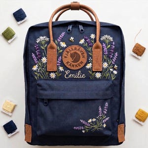 Op de afbeelding: Een marineblauwe rugzak met lichtbruine handvatten en een geborduurd bloemendesign met lavendel, witte madeliefjes en kleine bijen. De voorzijde toont een rond Fjällräven Kanken logo en de naam Emilie geborduurd in witte schrijfletters.
