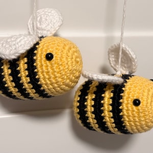 May include: Two crocheted bumblebees with black and yellow stripes and white wings. The bees are hanging from white strings.