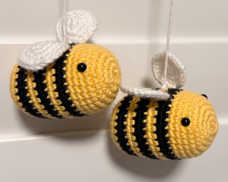 May include: Two crocheted bumblebees with black and yellow stripes and white wings. The bees are hanging from white strings.