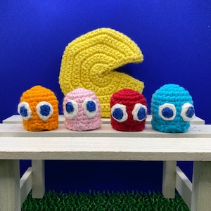 May include: Four crocheted Pac-Man ghosts in orange, pink, red, and blue, with white eyes and blue pupils, sitting on a white wooden bench in front of a yellow crocheted Pac-Man.