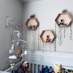 May include: A white crib with a white and gray mobile hanging above it. The mobile has a light blue balloon, white clouds, and yellow bees. There are three wooden hexagonal shelves on the wall behind the crib, each decorated with stuffed animals and greenery.