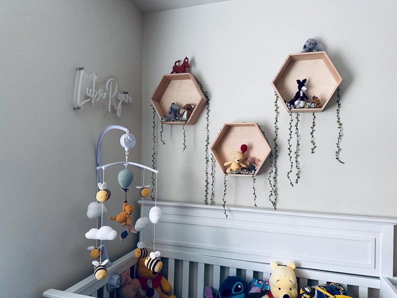 May include: A white crib with a white and gray mobile hanging above it. The mobile has a light blue balloon, white clouds, and yellow bees. There are three wooden hexagonal shelves on the wall behind the crib, each decorated with stuffed animals and greenery.