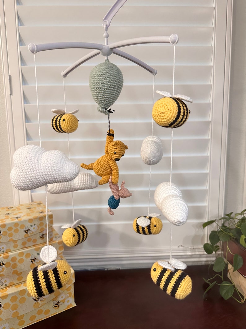 May include: A white baby mobile with a Winnie the Pooh and Piglet character, a green balloon, and six yellow and black bees. The mobile is hanging from a white ceiling.