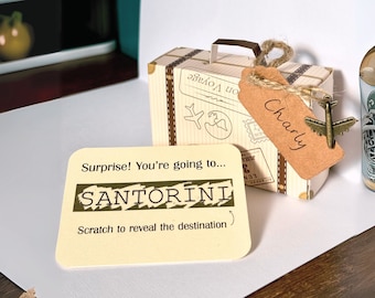 May include: A brown cardboard suitcase with a luggage tag that says "Charly" and a small airplane charm. The suitcase is sitting on a white surface with a card that says "Surprise! You're going to... SANTORINI Scratch to reveal the destination".