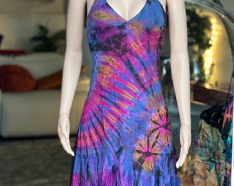Può includere: Abito all'americana tie-dye nelle tonalità del blu, viola e arancione. L'abito presenta uno scollo a V e un orlo svasato e asimmetrico. Il tessuto ha un motivo a vortice, con aree più scure che creano contrasto. L'abito è esposto su un manichino.